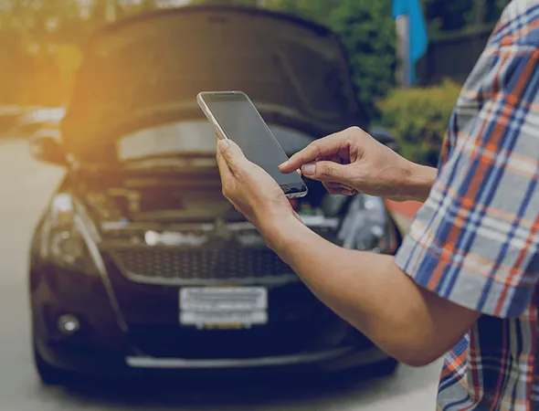 A person in a plaid shirt using a smartphone stands near a black car with the hood open, suggesting a breakdown. The scene conveys urgency.